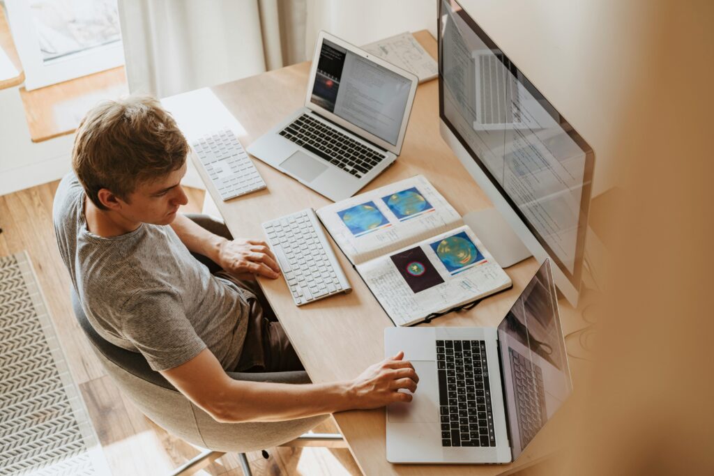 A man multitasks with laptops and a desktop, coding in a home office setting.